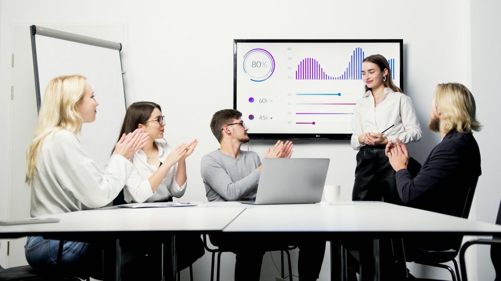 An image showing a woman displaying her dashboard and KPI's in a training scenario with people clapping. This resembles how governance and training are key to responsibly oversee the democratization of data visualization. 